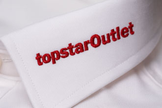 Gesticktes Logo auf Hemdkragen – Beispiel TopstarOutlet Stickerei für individuelle Firmenhemden Nahaufnahme eines weißen Hemdkragens mit aufgesticktem TopstarOutlet Logo in Rot – Logo Stickerei als Beispiel für personalisierte Business- und Firmenhemden
